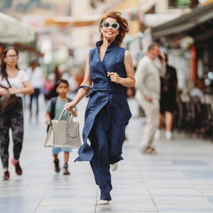 May include: A woman in a navy blue wrap jumpsuit and sunglasses runs down a city street, holding a light gray handbag. The outfit has a sleeveless top and wide-leg pants. Other people are walking in the background.