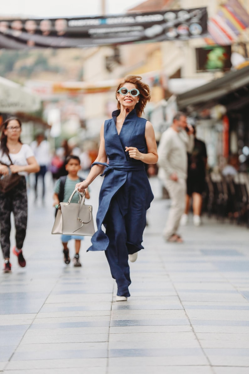 May include: A woman in a navy blue wrap jumpsuit and sunglasses runs down a city street, holding a light gray handbag. The outfit has a sleeveless top and wide-leg pants. Other people are walking in the background.