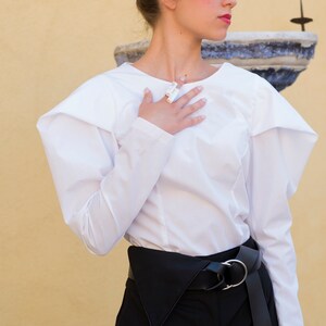 Puede incluir: Una blusa blanca de manga larga con hombros estructurados y pantalones negros de cintura alta. El atuendo se complementa con un cintur&oacute;n negro con hebilla plateada. La modelo lleva un anillo.