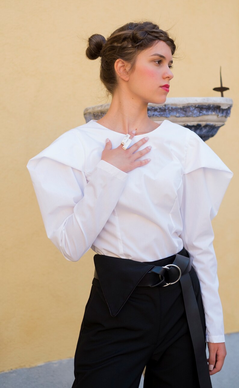 Puede incluir: Una blusa blanca de manga larga con hombros estructurados y pantalones negros de cintura alta. El atuendo se complementa con un cintur&oacute;n negro con hebilla plateada. La modelo lleva un anillo.