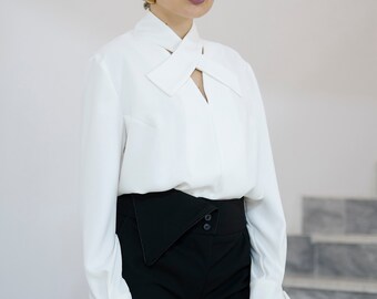 May include: A white long-sleeve blouse with a unique neckline and black high-waisted pants. The blouse features a distinctive cross-over design at the collar and cuffs. The pants have an asymmetrical waistline. The outfit is a modern take on classic styles.