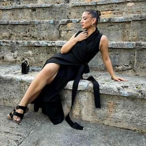 Puede incluir: Un vestido negro sin mangas con cintura envolvente y abertura alta. El vestido se combina con sandalias negras y un pequeño bolso negro. La persona está sentada en escalones de piedra.