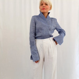 Puede incluir: Una blusa de lino azul claro con cuello mandarín y mangas acampanadas se combina con pantalones de lino blancos de pernera ancha. La blusa tiene un diseño cruzado. Los pantalones tienen cintura alta y pliegues.