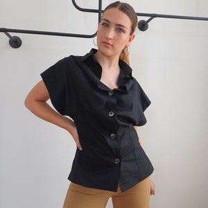 May include: Black short-sleeve button-down shirt with a tailored fit. The shirt features large, round buttons and a classic collar. The model is wearing tan cargo pants. A black metal clothing rack is in the background.