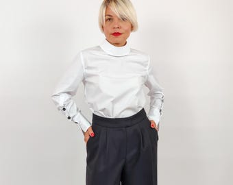 May include: A white long-sleeve blouse with a high collar and buttoned cuffs. The blouse is paired with black, wide-leg trousers. The buttons on the cuffs are black. The outfit is simple and elegant.