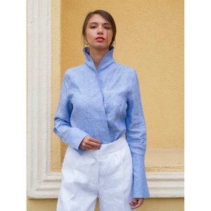 Può includere: Una camicia di lino azzurro con collo alto e maniche lunghe, abbinata a pantaloni di lino bianchi. La camicia presenta un design unico a portafoglio. L'outfit è ambientato su uno sfondo giallo.