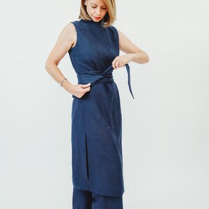 May include: A sleeveless, navy blue dress with a high neckline and a matching belt. The dress has a slit on the side and is paired with wide-leg pants. The model is adjusting the belt, showcasing the dress's design.