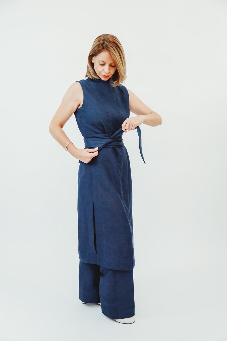May include: A sleeveless, navy blue dress with a high neckline and a matching belt. The dress has a slit on the side and is paired with wide-leg pants. The model is adjusting the belt, showcasing the dress's design.