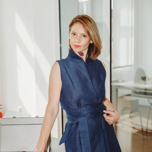 May include: A sleeveless, navy blue dress with a wrap-style bodice and a matching waist tie. The dress is worn by a person standing near a gray briefcase and a light gray table. The background includes a glass door and a bright, well-lit room.