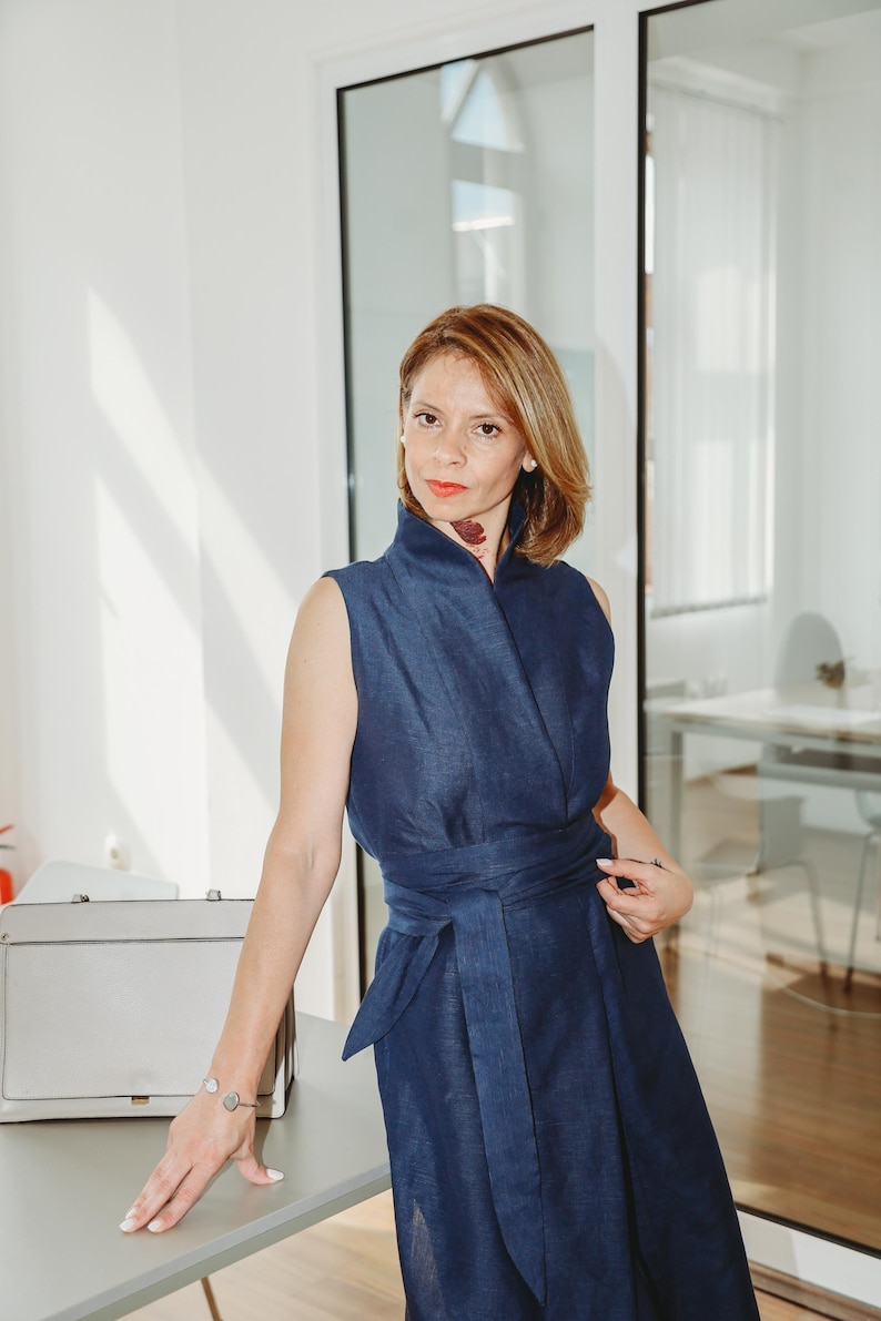 May include: A sleeveless, navy blue dress with a wrap-style bodice and a matching waist tie. The dress is worn by a person standing near a gray briefcase and a light gray table. The background includes a glass door and a bright, well-lit room.