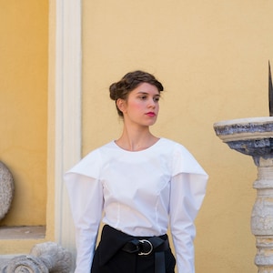 Puede incluir: Una mujer con una blusa blanca de manga larga con hombros estructurados y pantalones negros de cintura alta. El atuendo incluye un cintur&oacute;n negro con hebilla plateada. El fondo es una pared amarilla con detalles arquitect&oacute;nicos.
