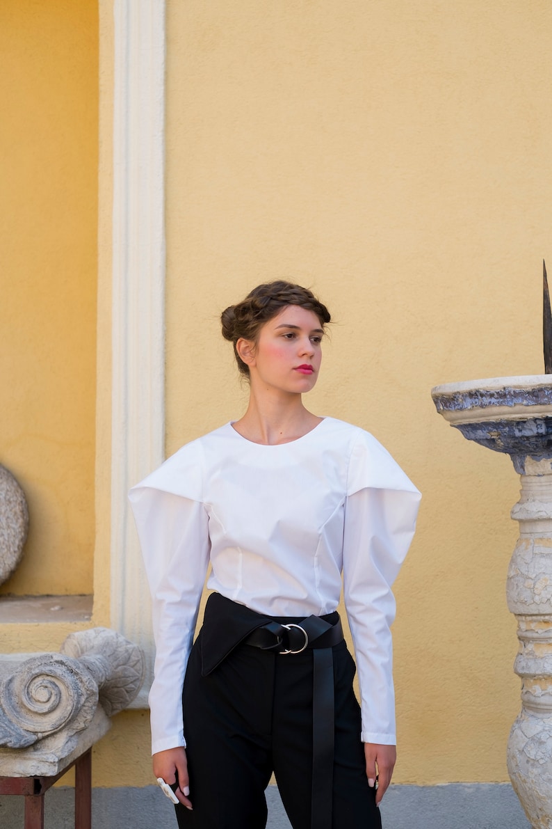 Puede incluir: Una mujer con una blusa blanca de manga larga con hombros estructurados y pantalones negros de cintura alta. El atuendo incluye un cintur&oacute;n negro con hebilla plateada. El fondo es una pared amarilla con detalles arquitect&oacute;nicos.