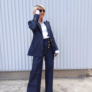 Könnte beinhalten: Ein dunkelblauer Jeansanzug mit Blazer und weitem Bein. Der Blazer hat goldene Knöpfe und wird über einem weißen Hemd mit Kragen getragen. Das Outfit wird durch goldene Sandalen und eine Sonnenbrille ergänzt.