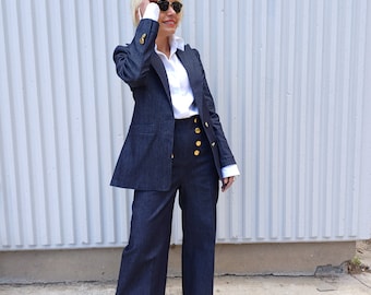 May include: A dark blue denim suit with a blazer and wide-leg pants. The blazer has gold buttons and a white collared shirt underneath. The outfit is paired with gold sandals and sunglasses. The suit is a classic, stylish ensemble.