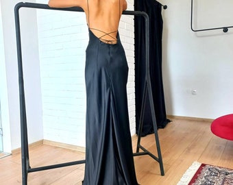 May include: A black satin evening gown with spaghetti straps and a low, crisscross back. The dress has a flowing skirt that pools on the floor. The model is leaning on a black metal clothing rack. The background includes a white brick wall and a black curtain.