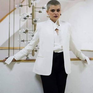 May include: A white blazer with a distinctive collar and long sleeves. The blazer is worn with a white blouse and black trousers. The sleeves have a cuff detail. The outfit is a modern take on a classic style.