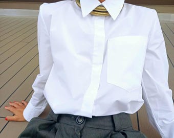 May include: A white shirt with a chest pocket and a gold-coloured collar necklace. The shirt is paired with dark grey pleated trousers. The outfit is simple and elegant.