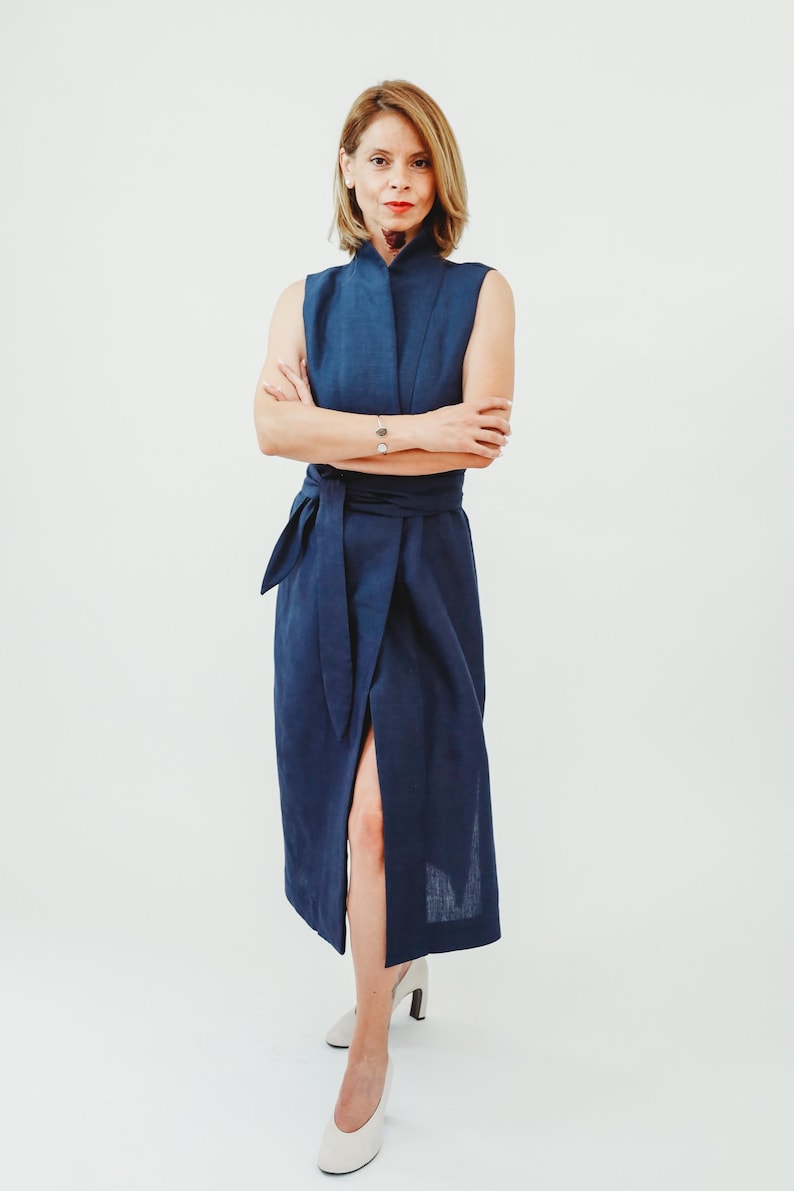 May include: A sleeveless, navy blue wrap dress with a high collar and a matching waist tie. The dress features a thigh-high slit and is paired with white heels. The model has short blonde hair and is standing in front of a white background.