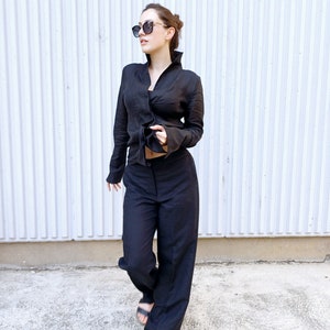May include: A black linen ensemble featuring a long-sleeved top with a high collar and matching wide-leg pants. The outfit is accessorized with sunglasses and sandals. The clothing is made of a lightweight fabric.