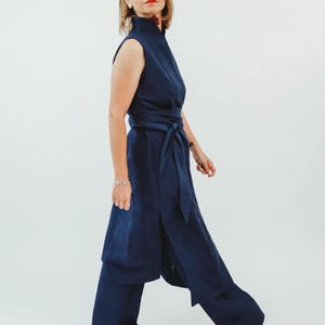 May include: A sleeveless navy blue jumpsuit with a high collar and a matching belt. The jumpsuit features wide-leg pants and is worn with white shoes. The outfit is made of a soft, flowing fabric, perfect for a stylish and comfortable look.
