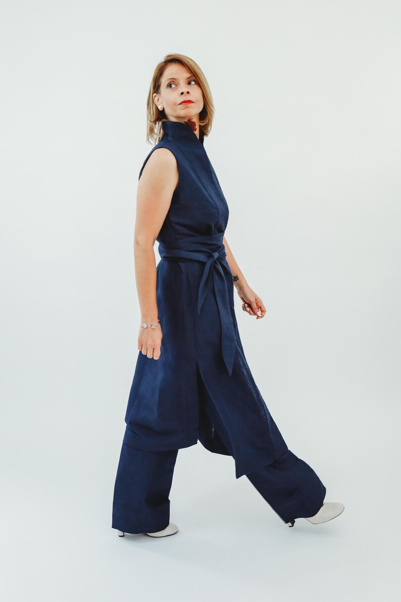 May include: A sleeveless navy blue jumpsuit with a high collar and a matching belt. The jumpsuit features wide-leg pants and is worn with white shoes. The outfit is made of a soft, flowing fabric, perfect for a stylish and comfortable look.