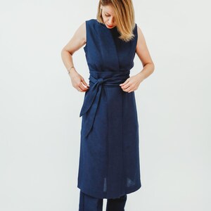 May include: A sleeveless navy blue dress with a matching belt tied at the waist. The dress is paired with navy blue pants. The model is wearing white shoes. The outfit is made of a soft, flowing fabric.