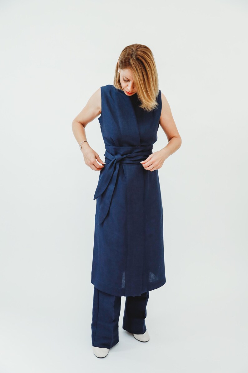 May include: A sleeveless navy blue dress with a matching belt tied at the waist. The dress is paired with navy blue pants. The model is wearing white shoes. The outfit is made of a soft, flowing fabric.