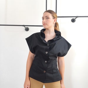 May include: Black button-down shirt with short, flared sleeves. The shirt has a fitted waist and a ruffled collar. The model is wearing tan cargo pants. The background is a plain white wall with black metal rods.