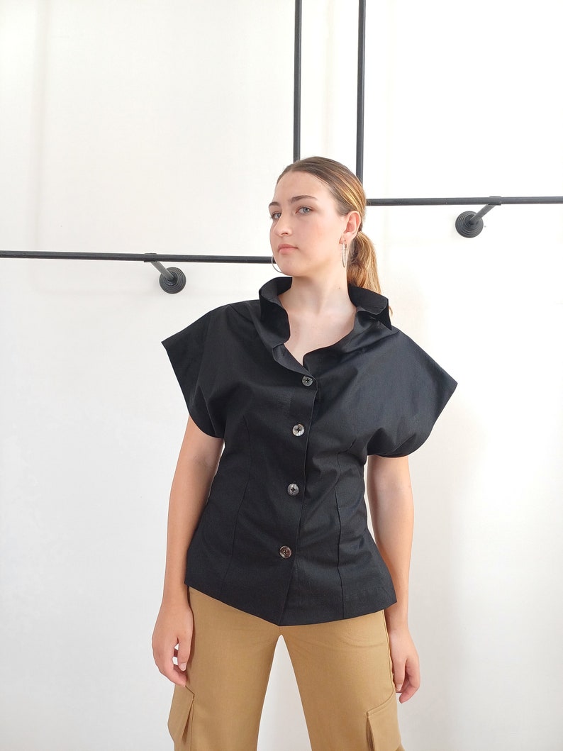 May include: Black button-down shirt with short, flared sleeves. The shirt has a fitted waist and a ruffled collar. The model is wearing tan cargo pants. The background is a plain white wall with black metal rods.