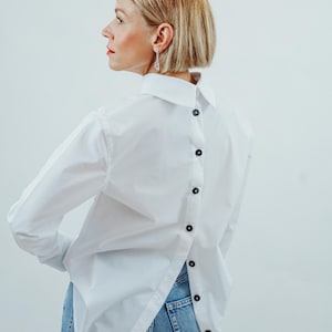 Könnte beinhalten: Ein weißes Hemd mit langen Ärmeln und einem einzigartigen Rückendesign. Das Hemd hat eine Reihe schwarzer Knöpfe auf der Rückseite und wird mit einer blauen Jeans kombiniert. Der Kragen ist klassisch.