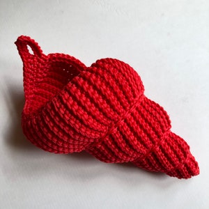 May include: A red crocheted seashell-shaped basket with a loop handle. The basket is made of thick, textured yarn and has a natural, organic look.