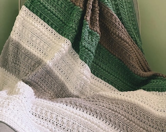 Könnte beinhalten: Eine handgefertigte Häkeldecke mit einem Farbblock-Design. Die Decke hat Streifen in Weiß, Grau, Grün und Braun. Der Häkelstich erzeugt eine strukturierte Oberfläche. Die Decke ist über einen grauen Stuhl drapiert.