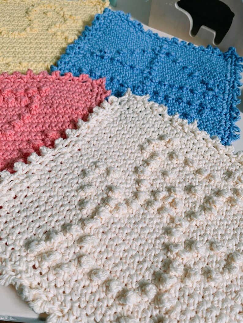 Puede incluir: Cuatro pa&ntilde;os de cocina de ganchillo en diferentes colores: amarillo, azul, rosa y blanco. Los pa&ntilde;os de cocina tienen una superficie texturizada y abultada.