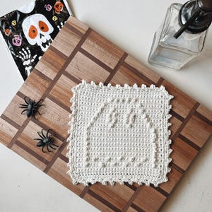 May include: A cream-colored, crocheted square with a house design, placed on a wooden cutting board. Two plastic spiders and a Halloween-themed bag with a skull graphic are also present. A glass soap dispenser is in the background.