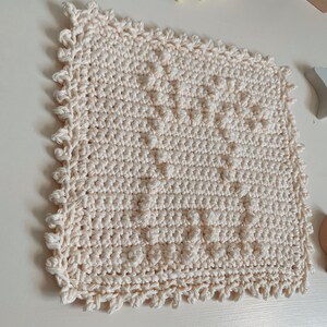 Puede incluir: Un pa&ntilde;o de cocina cuadrado de color crema tejido a crochet con un patr&oacute;n texturizado y un borde festoneado.