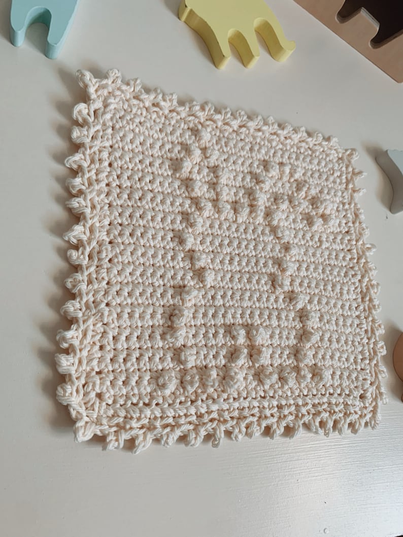 Puede incluir: Un pa&ntilde;o de cocina cuadrado de color crema tejido a crochet con un patr&oacute;n texturizado y un borde festoneado.