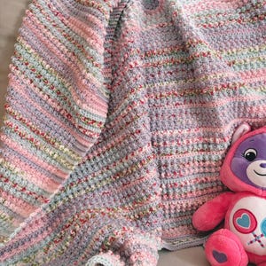 May include: A handmade, pastel-colored crochet blanket with pink, blue, and lavender stripes. The blanket has a textured, bobble stitch design and is draped over a surface. A pink stuffed bear with a purple face is next to the blanket.