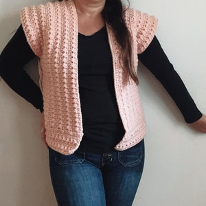 May include: A pink crocheted vest with a V-neck and short sleeves. The vest is worn over a black shirt and blue jeans.