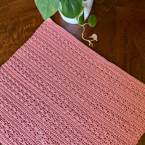 May include: A pink crocheted blanket with a textured pattern. The blanket is lying on a wooden surface.