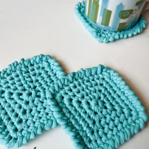May include: Two teal blue crocheted coasters. The coasters are square shaped with a textured design.