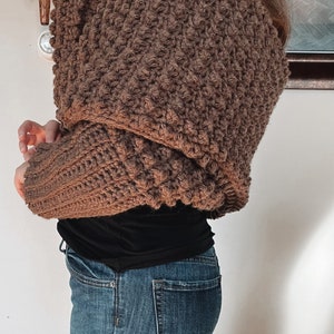 May include: A brown crocheted shrug with a textured stitch pattern. The shrug is worn over a black shirt and blue jeans.