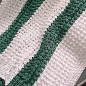 May include: A handmade white and green knitted blanket. The blanket features alternating stripes of white and green, with a textured, raised stitch pattern. The green stripes have a unique, bobble-like design, adding visual interest to the cozy textile.