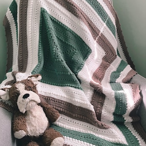 May include: A handmade crochet blanket in shades of white, green, and brown, draped over a gray chair. The blanket features a diagonal striped pattern. A stuffed dog toy with brown and white fur rests on the chair.
