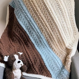 Könnte beinhalten: Eine gehäkelte Babydecke mit einem diagonalen Muster aus braunen, blauen, beigen und weißen Streifen. Die Decke hat eine strukturierte, noppenartige Oberfläche.