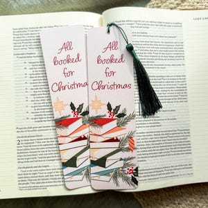 May include: Two rectangular bookmarks with the text "All booked for Christmas" in red script. The bookmarks feature illustrations of stacked books, holly, and stars. A green tassel hangs from one bookmark. The bookmarks are placed on an open book.