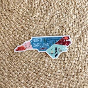May include: A white sticker with a colorful illustration of the state of North Carolina. The sticker features various landmarks and symbols, including a lighthouse, a mountain range, and a beach scene. The text "NORTH CAROLINA" is printed in black at the top of the sticker.
