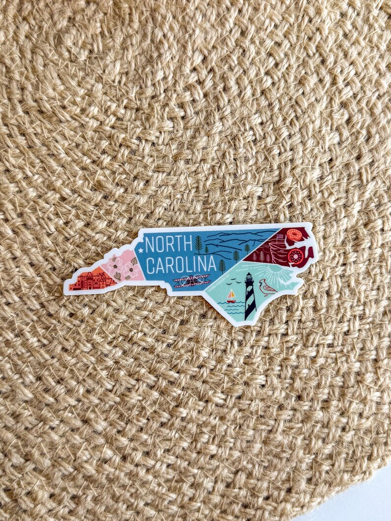 May include: A white sticker with a colorful illustration of the state of North Carolina. The sticker features various landmarks and symbols, including a lighthouse, a mountain range, and a beach scene. The text "NORTH CAROLINA" is printed in black at the top of the sticker.