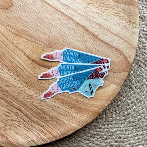 May include: Three North Carolina state outline stickers with a colorful design. The stickers are stacked on top of each other on a wooden surface.