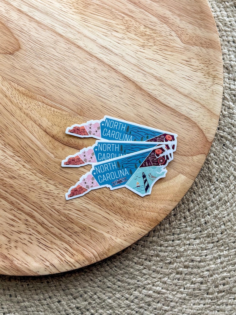 May include: Three North Carolina state outline stickers with a colorful design. The stickers are stacked on top of each other on a wooden surface.