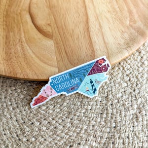May include: A white sticker with a colorful outline of the state of North Carolina. The sticker features various illustrations of landmarks and natural features, including a lighthouse, mountains, and a beach scene. The text "NORTH CAROLINA" is printed in black letters on the sticker.